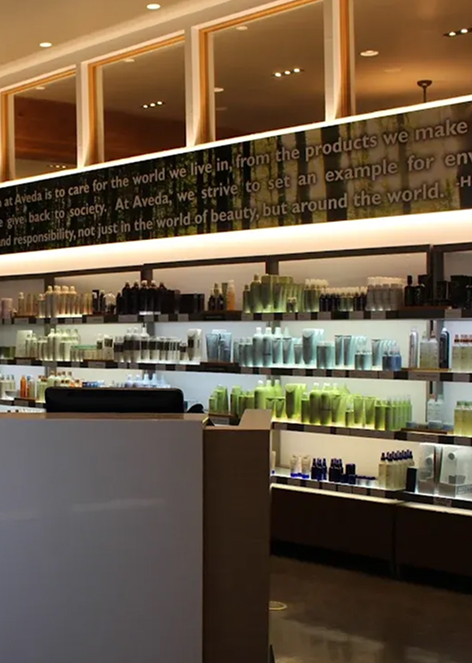 - Indira Salon Spa in Waupaca, WI Modern salon interior with shelves displaying various hair and beauty products, illuminated by overhead and backlighting. - Indira Salon Spa in Waupaca, WI