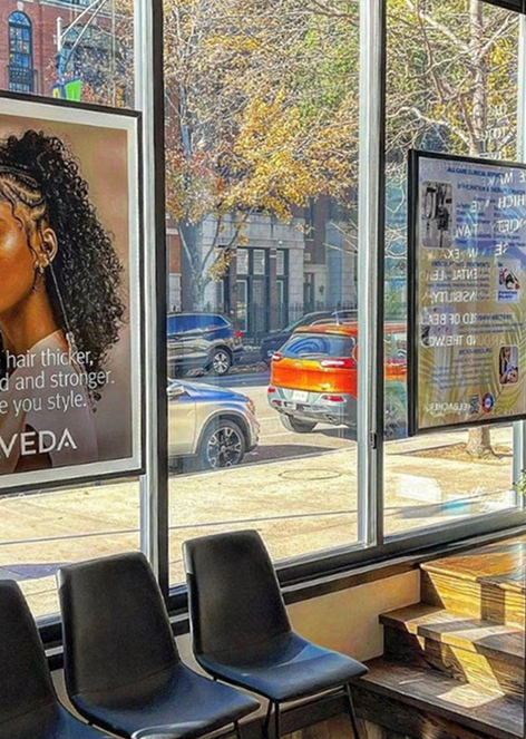 - Indira Salon Spa in Waupaca, WI Row of black chairs by large windows inside a salon or waiting area, with posters on the glass and cars visible parked outside on a sunny day. - Indira Salon Spa in Waupaca, WI