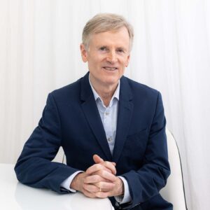 A person in a blue blazer and light shirt sits at a table with hands clasped, perhaps contemplating the benefits of Ultraceuticals, all set against a white curtain background. | Indira Salon Spa