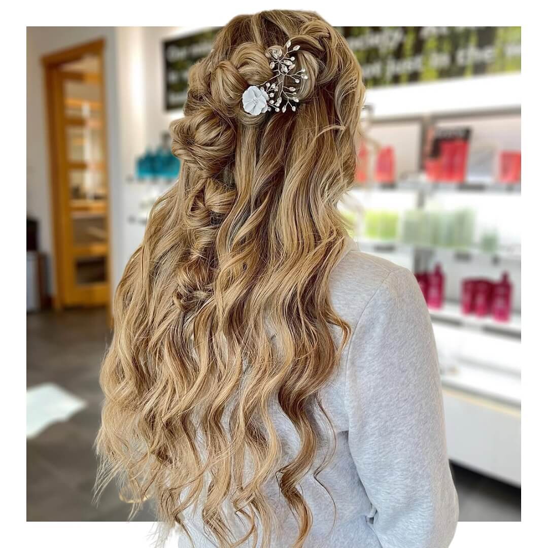 - Indira Salon Spa in Chicago IL, Park Ridge IL, Green Bay WI A woman with long, wavy blonde hair styled in loose curls and a half-up braid, decorated with a jeweled hair accessory, stands in a modern salon. - Indira Salon Spa in Chicago IL, Park Ridge IL, Green Bay WI