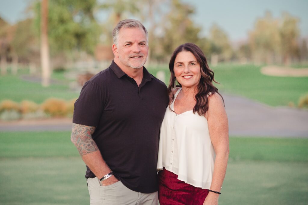 A man and a woman stand together outdoors on a grassy area, smiling at the camera with trees and a path in the background, radiating wellness and natural beauty. - Indira Salon Spa in Chicago IL, Park Ridge IL, Green Bay WI